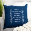 A navy blue throw pillow on a gray sofa with white floral designs and text that reads: "DON'T CROSS YOUR FINGERS AND HOPE. FOLD YOUR HANDS AND PRAY." A white blanket and green plant are in the background.