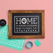 A framed chalkboard sign on a pink surface reads "Home is where someone runs to greet you" with a paw print in the word "home." Beside it are chalk paste, a stencil, and an open paint container.
