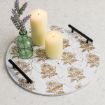 A round serving tray with black handles features a white and gold floral pattern. On the tray are two lit pillar candles and a small green glass bottle holding sprigs of lavender.