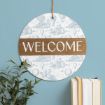 A round sign with a white and light blue pattern, a brown stripe in the center displaying the word "WELCOME" in white letters, hangs on a wall above a plant and a stack of books.