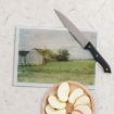 A kitchen scene with a cutting board featuring a rural barn painting, a black-handled knife resting on it, and a round wooden plate with sliced apples on a light countertop.