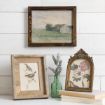 A rustic wall displays a landscape painting and rests two framed artworks—one of birds, one of flowers—on a stack of books and a green glass vase with lavender sprigs.