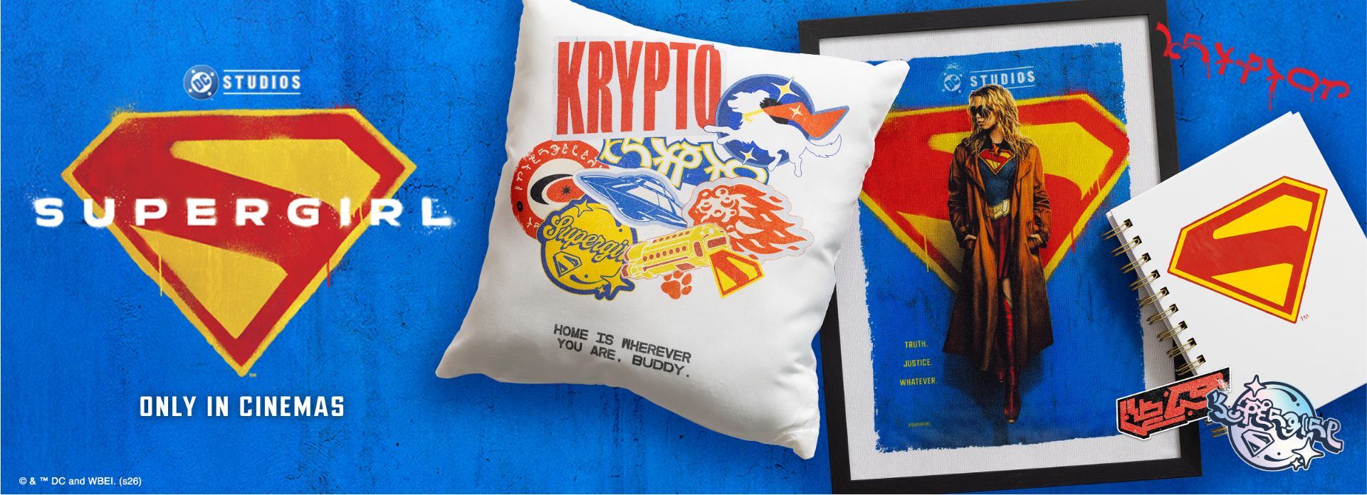 A display of Supergirl-themed merchandise includes a Supergirl logo, a pillow with "Krypto" and graffiti designs, a framed Supergirl poster, a notebook with the logo, and stickers. Text: "SUPERGIRL ONLY IN CINEMAS.