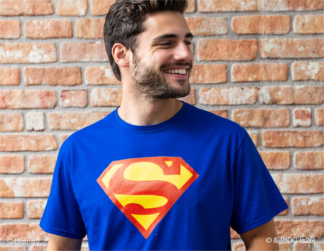 A smiling man with short brown hair and a beard wears a bright blue T-shirt featuring the red and yellow Superman “S” logo, standing in front of a brick wall.