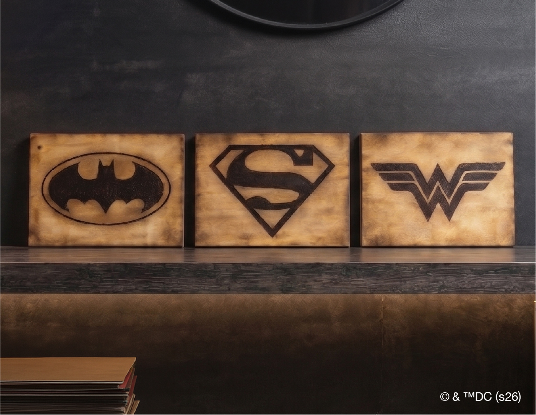 Three wooden plaques display the Batman, Superman, and Wonder Woman logos, arranged side by side on a shelf against a dark wall.