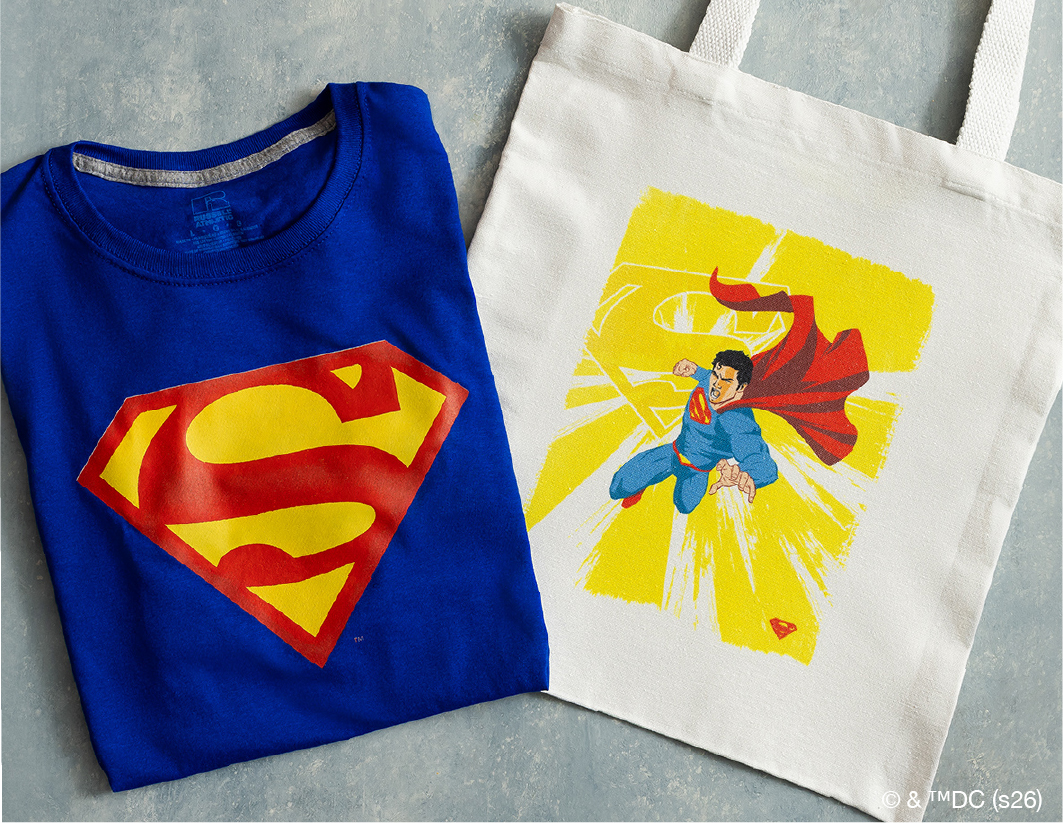 A blue T-shirt with a large yellow and red Superman logo next to a white tote bag featuring an illustration of Superman flying with his cape flowing and a bright yellow background.