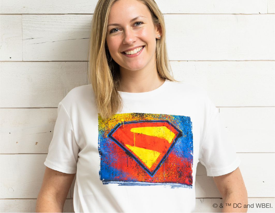 A smiling woman with blonde hair wears a white t-shirt featuring a colorful, artistic Superman logo design, standing against a white wooden wall.