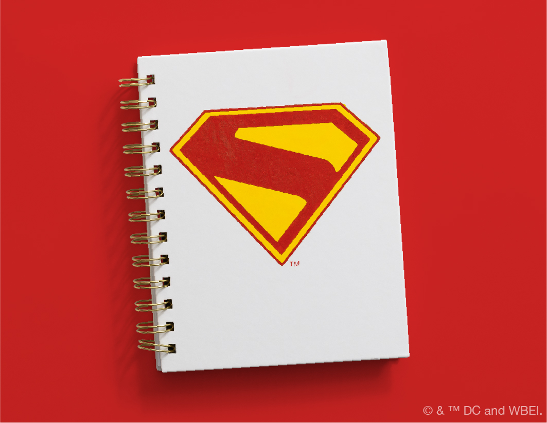 A white spiral-bound notebook with a yellow and red Superman logo on the cover, placed on a solid red background. The DC and WBIE copyright notice is visible in the bottom right corner.