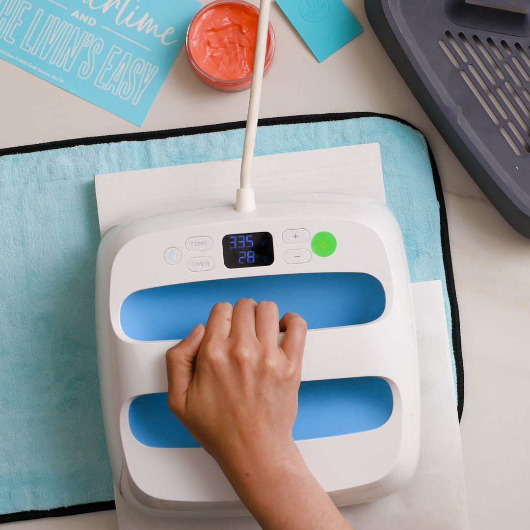 A hand presses down on a white and blue heat press machine, set to 335°F for 28 seconds, on a piece of white material over a light blue mat. Craft supplies are visible around the work area.