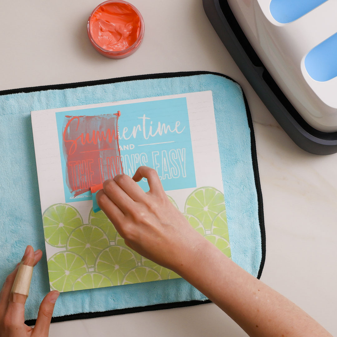 A person uses a squeegee to apply red paint over a stencil that says “Summertime and the livin’s easy” on a blue and green design with lime slices, on a white surface with a towel underneath.