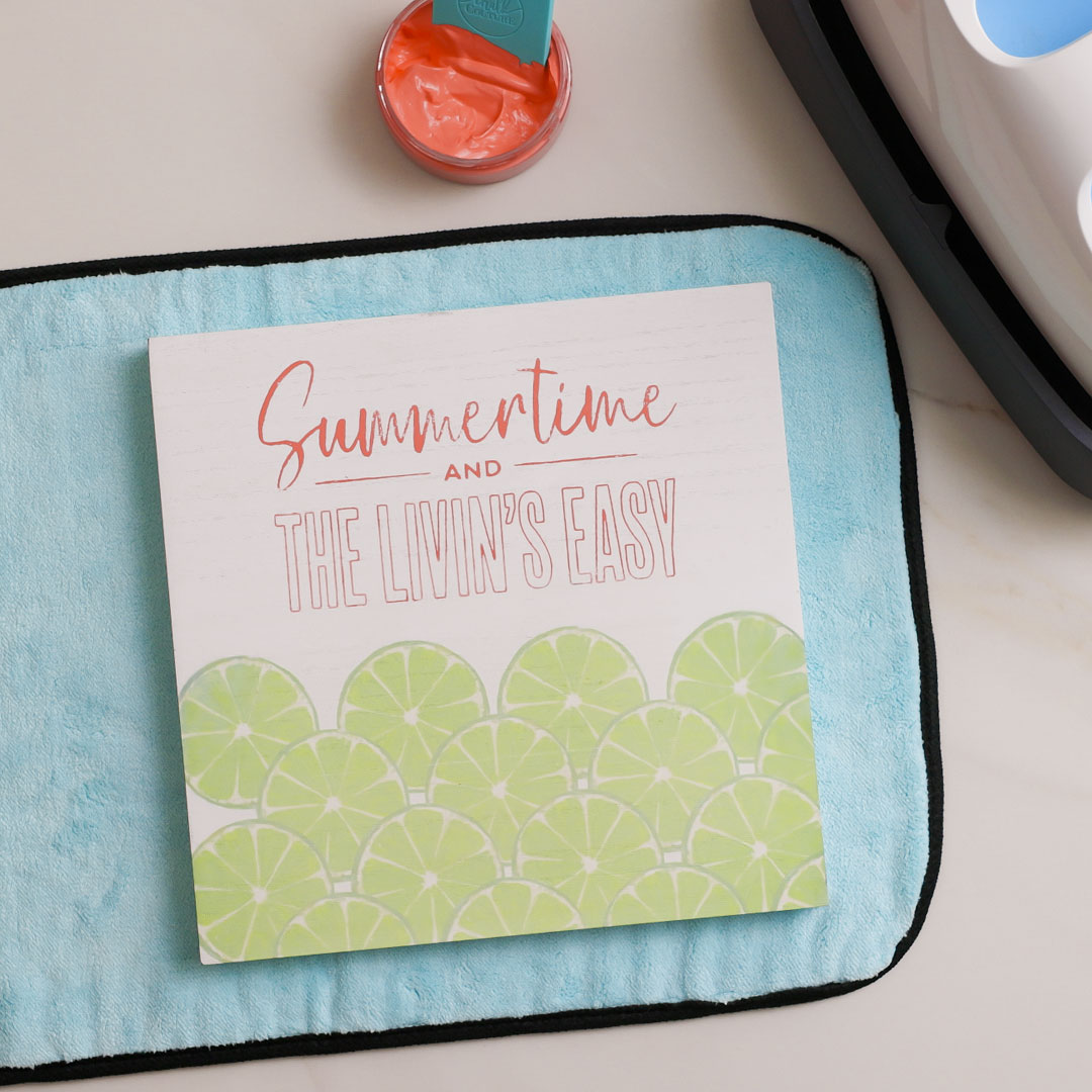 A square sign with lime slice illustrations and the words "Summertime and the Livin’s Easy" in orange and teal lettering, placed on a light blue cloth with a small jar of orange paint nearby.