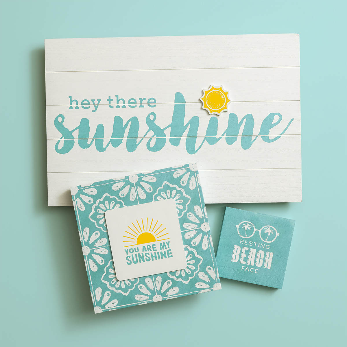 A decorative sign reads "hey there sunshine" in teal and white with a sun icon. Below are two coasters: one says "You are my sunshine" with a sun, the other says "Resting beach face" in white on teal.