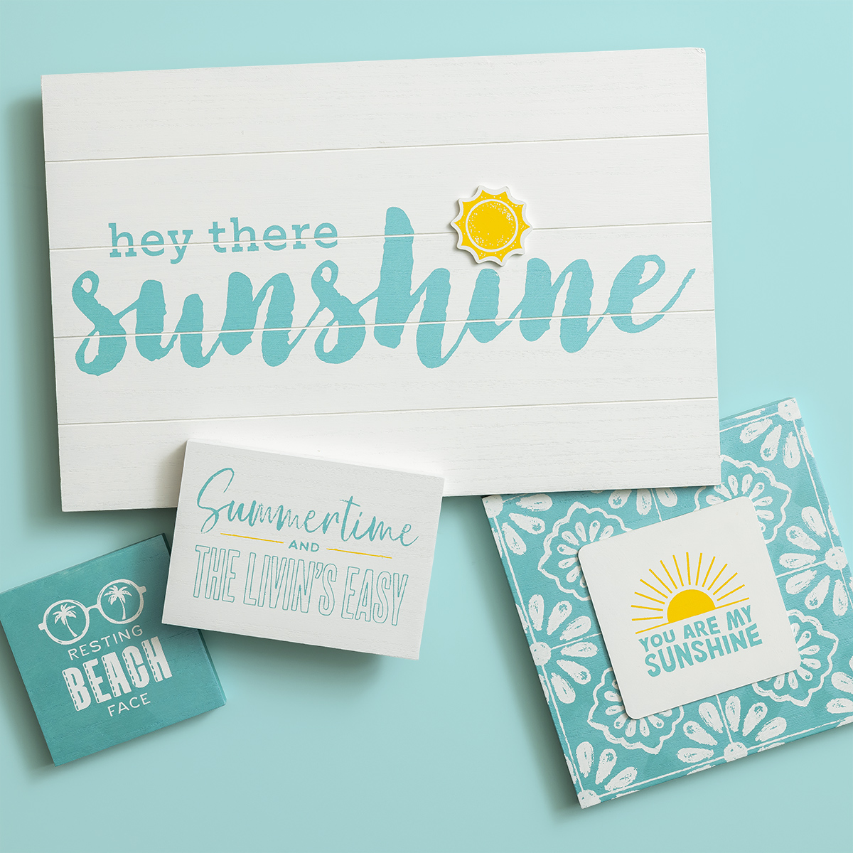 A flat lay of four summer-themed signs on a light blue background. The largest says "hey there sunshine" with a sun icon. The others read "Resting Beach Face," "Summertime and the Livin's Easy," and "You Are My Sunshine.