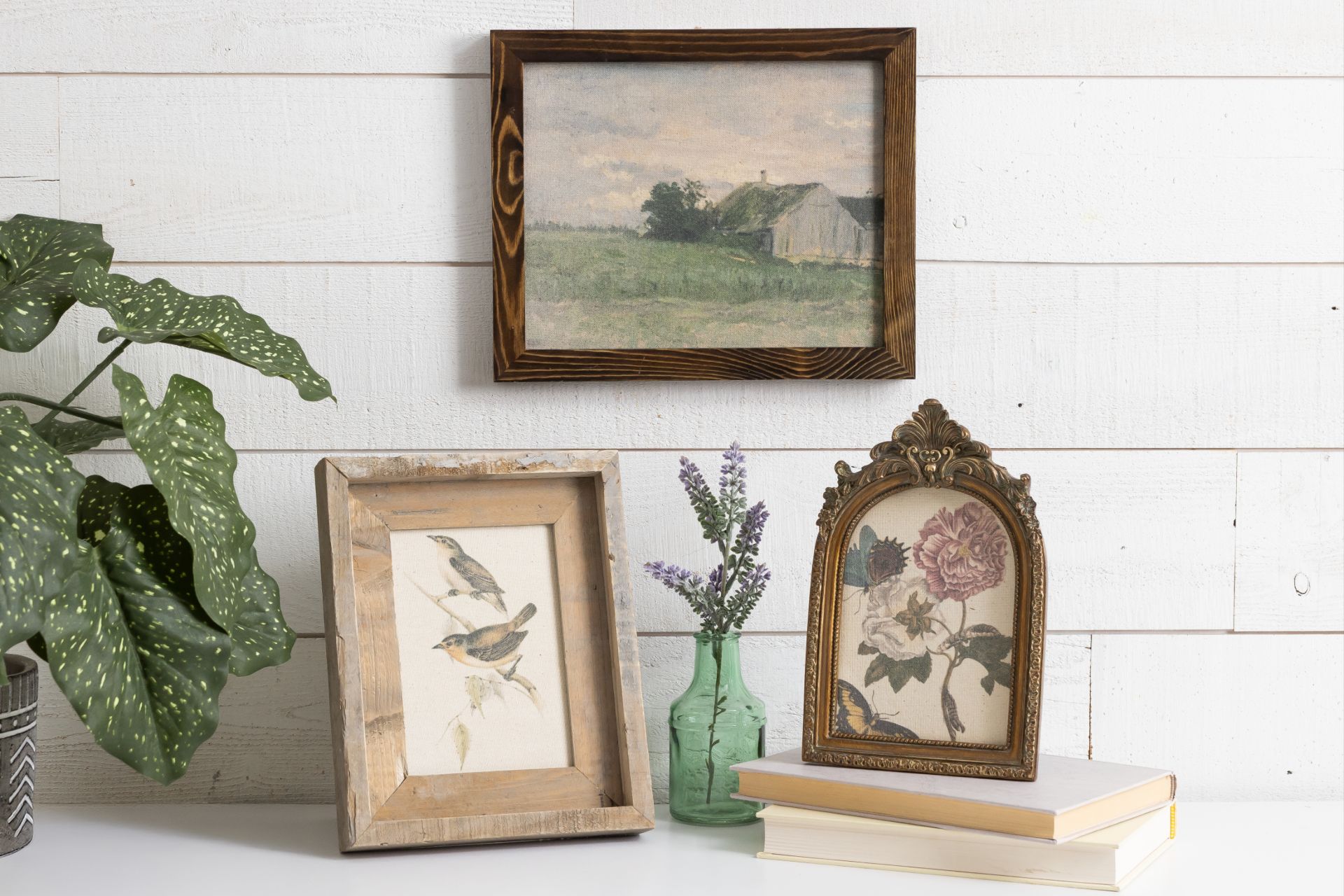 A white tabletop with a potted green plant, two framed floral and bird illustrations, books, and a small vase with lavender. A rustic landscape painting hangs on a white shiplap wall in the background.
