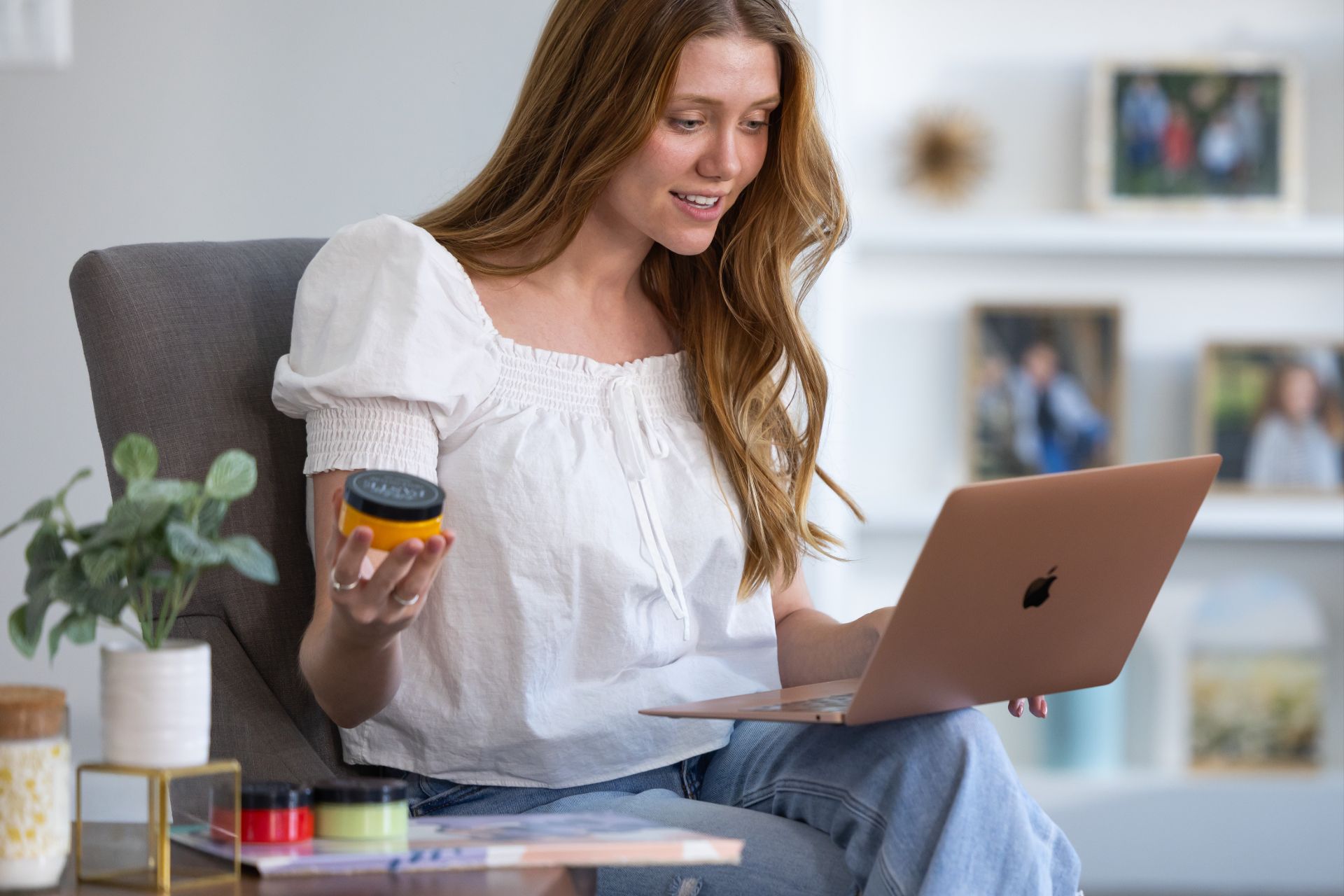 A woman sits in a chair holding a small round container in one hand and looking at a laptop on her lap, smiling. She is indoors with photos and a plant in the background.