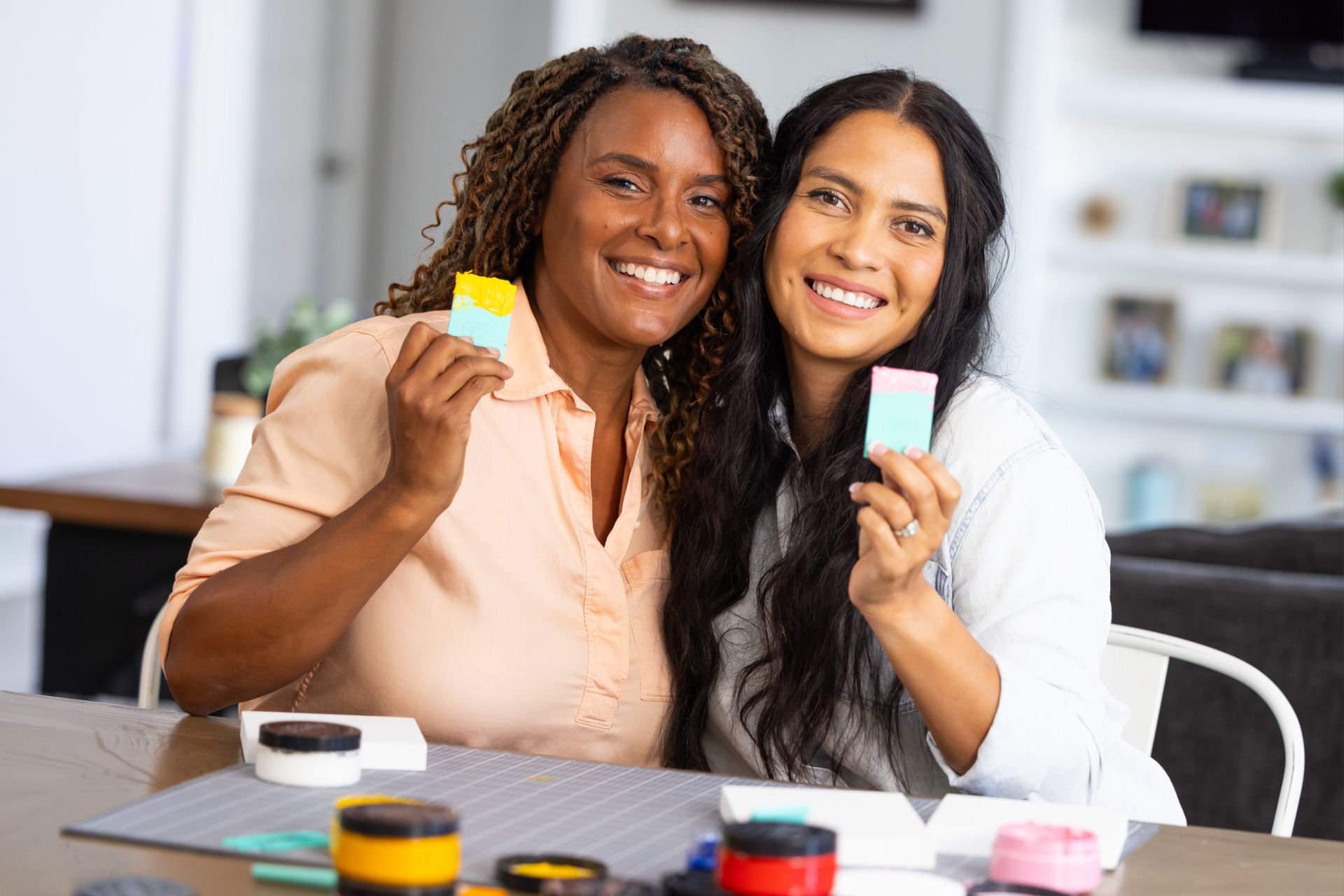Two women smiling and sitting at a table, holding up colorful handmade soap bars. Craft supplies are spread out in front of them, and they appear happy and engaged in a creative activity at home.