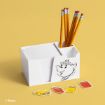 A white desk organizer with paper and pencils features a drawing of Mrs. Potts from Disney’s Beauty and the Beast. Four colorful keychains shaped like books and a pencil are placed in front on a yellow background.