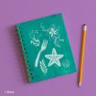 A teal spiral notebook with white sketches of a fork, seaweed, starfish, seashell, Ariel, and a flower, placed on a purple surface next to a yellow pencil. Disney logo is in the corner.