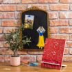 A decorative desk setup features a potted plant, red book stand with white apple and flower designs, a framed blackboard with a Snow White illustration, a photo, and office supplies, all against a brick wall.