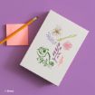 A white notebook with colorful illustrations of a chameleon, flowers, and leaves on the cover sits on a purple surface next to a pencil and a pink sticky note. Disney logo is visible in the corner.