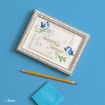 A framed card with blue birds, flowers, and the gold words "happily, forever" lies on a blue surface beside a pencil and a blue sticky note. Disney logo is visible in the corner.