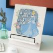 A wooden plaque with an illustration of Cinderella, flowers, and the words “Dreams is a wish your heart makes” displayed on a white stand beside a glass vase with greenery on a desk.