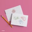 A greeting card on a pink surface features colorful line drawings of Disney’s three Sleeping Beauty fairies and small stars, with a matching envelope decorated with a rose illustration. A pencil lies nearby.