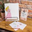 A wooden desk with a drawing of Princess Aurora on a stand, colored sketches on white paper, a gold pen, a clear tape dispenser, and a pink framed sign that reads “Believe in Yourself” near a brick wall.