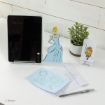A tablet on a stand is next to a Cinderella drawing, a potted plant, a small phone stand with Lumière from Beauty and the Beast, and blue and white papers with pencil sketches of Cinderella-themed designs. A pen lies nearby.