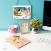 A bright desk setup with a framed drawing and a potted plant, a mug, a keyboard, a pencil, and a notepad featuring Moana. Above is a wall hanging showing an ocean scene and a sticky note with hearts.