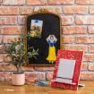 A desk with a small potted plant, stationery, and an open red notebook. Behind them, a decorative chalkboard displays a photo, a cutout of Snow White, and hangs on a brick wall. © Disney.