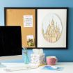 A desk with a computer, a pink mug, a tissue box, and a small vase sits in front of a corkboard. The board has a weekly planner, a photo, and a colorful castle illustration displayed.