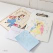 A Disney-themed notebook, a clipboard with Disney princesses, a white envelope, and a light blue sheet with decorative sketches are arranged on a white surface.