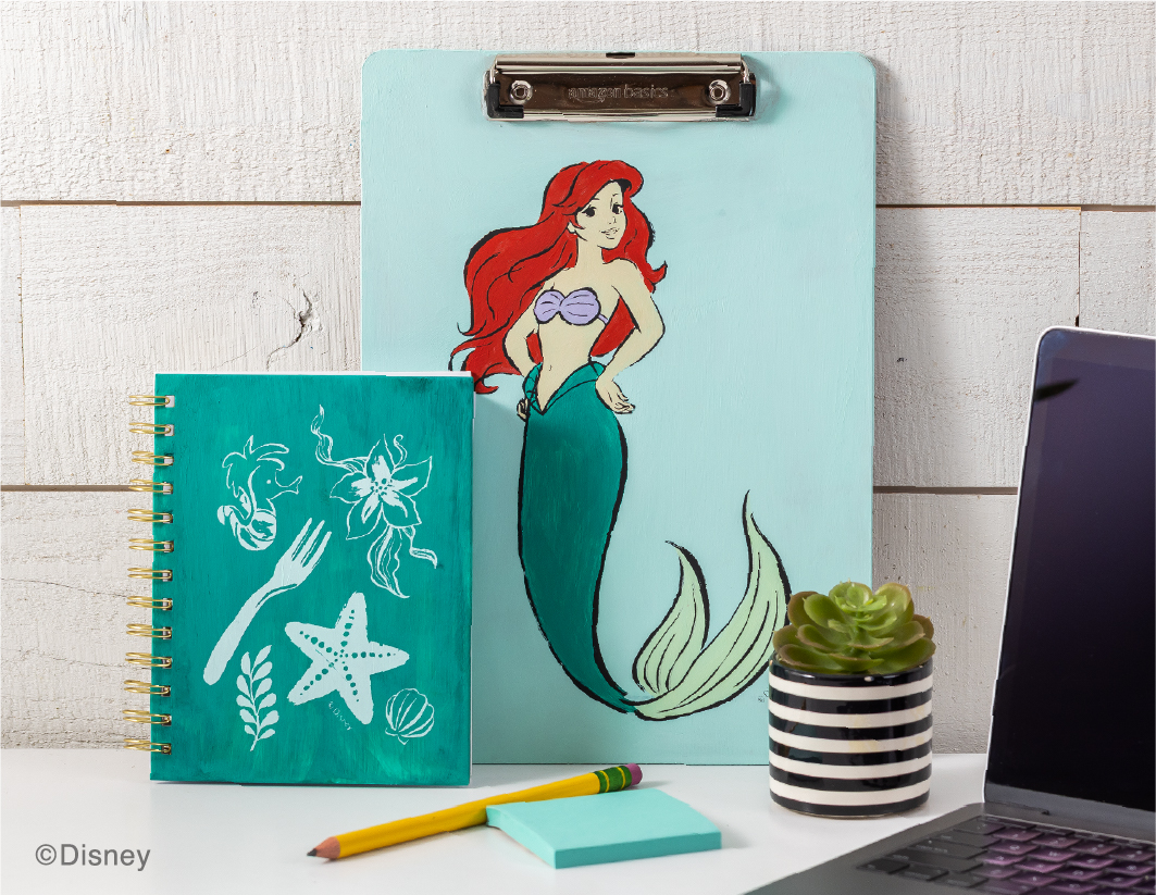 A painted clipboard with Ariel from "The Little Mermaid" stands behind a teal notebook with white sea designs. A pencil, sticky notes, and a small striped potted succulent are on the desk beside a laptop.