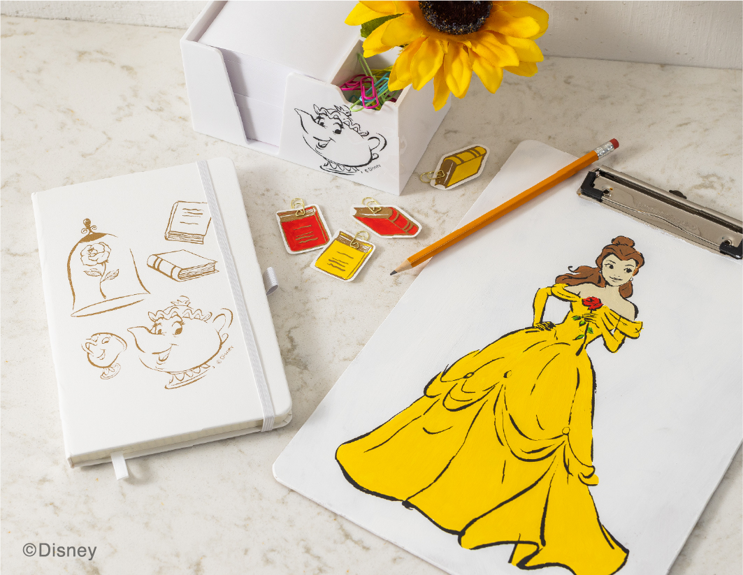 A drawing of Belle in her yellow dress is on a clipboard beside a pencil. Nearby are Beauty and the Beast-themed charms, a white notebook with sketches, a sunflower, and a box with more Disney-themed notes. ©Disney.