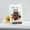 A decorative card on a stand features an illustration of a lit candle in a jar, cinnamon sticks, orange slices, and autumn berries with the text "autumn living." A beige flower sits nearby on a white surface.