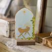 A decorative glass plaque on a wooden stand features a gold illustration of baby Jesus in a manger, with a golden star above and green leafy accents on the side. The plaque is displayed on a lace-covered table.