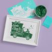 A framed print of Santa Claus driving a truck with "Santa Claus is coming to town!" in green ink, next to a stencil, a tin of green paste, and a teal card on a purple surface.