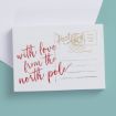 A postcard with "with love from the north pole" written in red script, and gold "Post Card" and postage details in the top right corner, placed on a light blue surface with part of a white envelope showing.