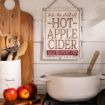 A kitchen scene with a pot on the stove, apples on the counter, a container of utensils, and a vintage-style sign on the wall that reads, "Take the chill off HOT APPLE CIDER.