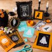 A collection of Halloween-themed crafts and decorations, including illustrated cards, treat bags, scissors, candles, candies, and framed artwork featuring ghosts, a pumpkin, a witch hat, and a black cat on a wooden table.
