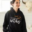 A smiling woman wearing a black hoodie with the text "It's October witches" and bat graphics, standing indoors with a white wall and a decorative animal skull in the background.