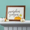 A framed sign with the words "pumpkin season" and an illustration of a pumpkin sits on a white mantel with three small decorative pumpkins in front. The wall behind is painted light teal.