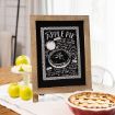 A framed chalkboard apple pie recipe drawing stands on a table next to a lattice-topped apple pie and a cake stand holding green apples. Sunlight filters through sheer curtains in the background.