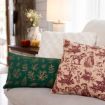 Two decorative pillows on a beige couch: one green with gold leaves, the other cream with maroon vintage-style animal and people illustrations. Soft lighting, a white lamp, and small pumpkins are in the background.