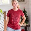 A smiling woman wearing a red T-shirt with a festive design and “Merry Christmas” text stands indoors near a decorated Christmas tree.