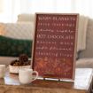 A framed sign on a wooden table lists cozy autumn phrases, with pinecones and two white mugs in front. A green pillow and cream blanket are on a couch in the background. Warm light fills the room.