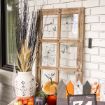 A rustic wooden window frame decorated with fake spider webs, surrounded by a white vase holding twigs, assorted pumpkins, and wooden decorations in the shape of houses, creating a Halloween-themed display.