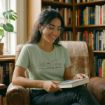 A smiling woman with glasses sits in a cozy armchair, holding an open book. She wears a light green shirt that says "book reading season," and is surrounded by bookshelves filled with books.