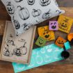 A wooden table with Halloween crafts: a pillow and framed art with pumpkin sketches, painted wood blocks with jack-o'-lantern faces, paint jars, and a stencil sheet with pumpkin and face designs.